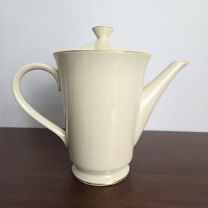 May include: Cream-colored ceramic teapot with a lid and a curved handle. The teapot has a spout and is trimmed with gold accents. The teapot is on a dark brown surface.