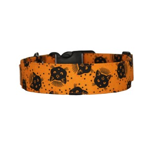 May include: Orange dog collar with a black cat face pattern. The collar has a black buckle and is made of fabric.