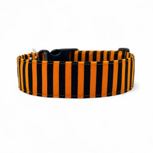 Może przedstawiać: Obroża dla psa z pionowymi pomarańczowo-czarnymi paskami. Obroża posiada czarną plastikową klamrę i srebrną metalową pętlę do przypięcia smyczy. Wykonana z wytrzymałej tkaniny.