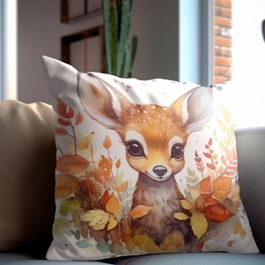 May include: A white throw pillow with a watercolor illustration of a fawn with brown and white spots surrounded by fall leaves in shades of orange, yellow, and brown.