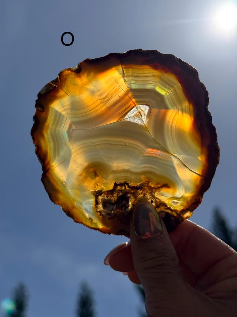 May include: A polished agate slice, displaying bands of amber, gold, and brown. The natural stone is held up to the sunlight, revealing its intricate patterns and textures. The agate's edges are rough, and the overall shape is irregular. The agate is a natural stone.