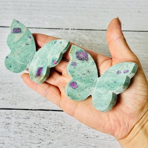 May include: Two butterfly-shaped carvings made of green fuchsite stone with embedded magenta ruby inclusions. Each piece features a smooth, flat surface and distinct wing contours, showcasing the natural color contrast between the light green base and the vibrant purple-red mineral spots.