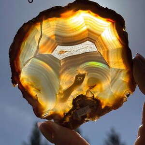 May include: A layered agate stone with translucent bands of orange, brown, and white. The stone is held up to the light, showcasing its intricate patterns and natural beauty. The text "J" is in the upper left corner.