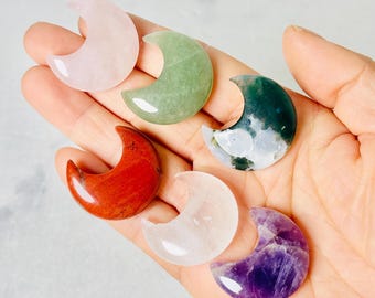 May include: Six polished crescent moon-shaped gemstones in various colors: pink, green, red, clear, and purple. The gemstones are held in a hand, showcasing their smooth, reflective surfaces. The gemstones are likely used for decorative or spiritual purposes.