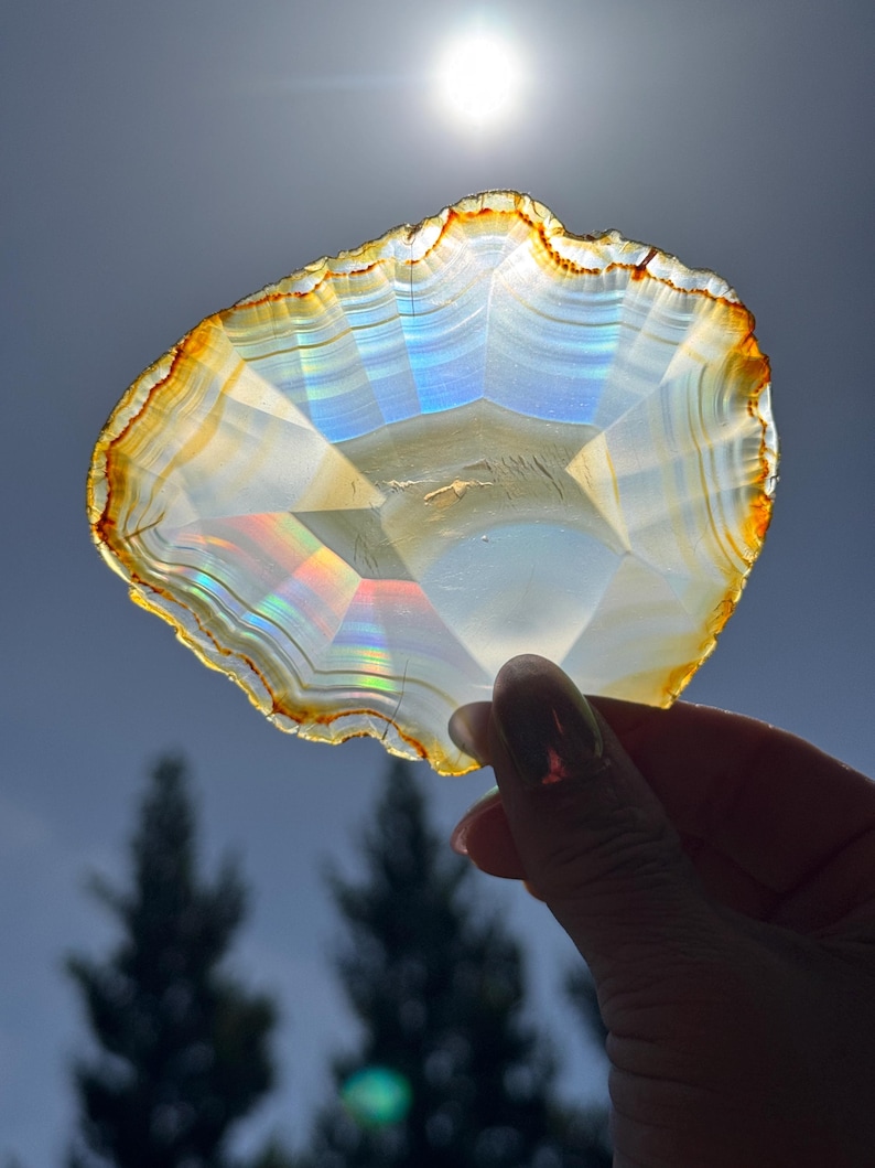 May include: A hand holds a translucent agate slice, showcasing natural bands of white, yellow, and orange. The polished stone has an irregular edge and internal rainbow iridescence. The light shines through the stone against a bright sky.