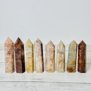 May include: Seven natural stone obelisks in varying shades of brown, tan, and cream are arranged in a row. Each obelisk has a rectangular base and tapers to a pointed top. The stones have unique patterns and textures.
