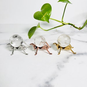 May include: Three clear quartz crystal spheres on decorative metal stands. The stands are silver, rose gold, and gold.