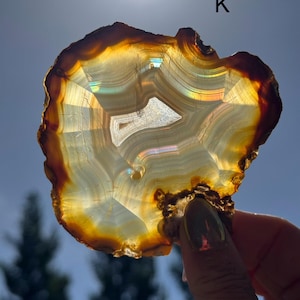 May include: A hand holds a polished agate slice, showcasing natural bands of brown, gold, and white. The translucent stone has an organic shape and a lighter central area. The letter "K" is visible in the background.
