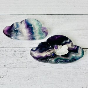 May include: Two cloud-shaped trays made of polished fluorite. The trays are primarily purple, with streaks of blue, green, and white. One tray holds a silver ring with a blue heart-shaped stone and a small crystal.