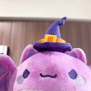 May include: Purple plush bat with a purple witch hat and orange trim. The bat has embroidered eyes and a small mouth.