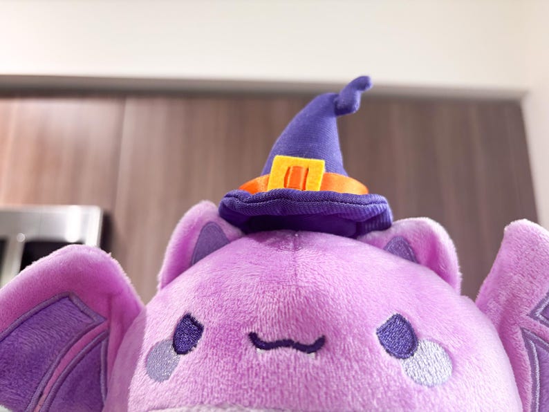 May include: Purple plush bat with a purple witch hat and orange trim. The bat has embroidered eyes and a small mouth.