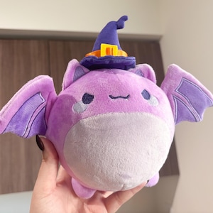 May include: A plush purple bat wearing a purple and orange witch hat with a yellow buckle. The bat has a round body and large, purple wings.