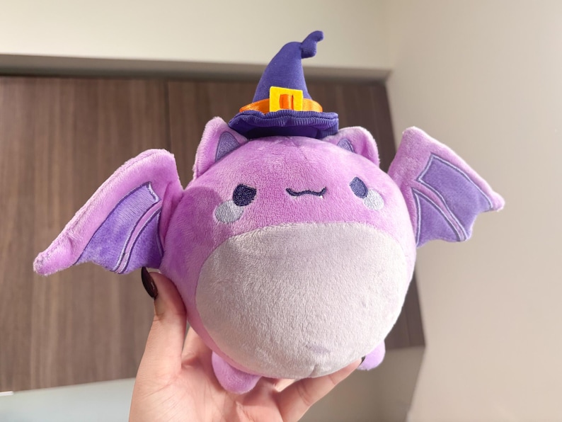 May include: A plush purple bat wearing a purple and orange witch hat with a yellow buckle. The bat has a round body and large, purple wings.