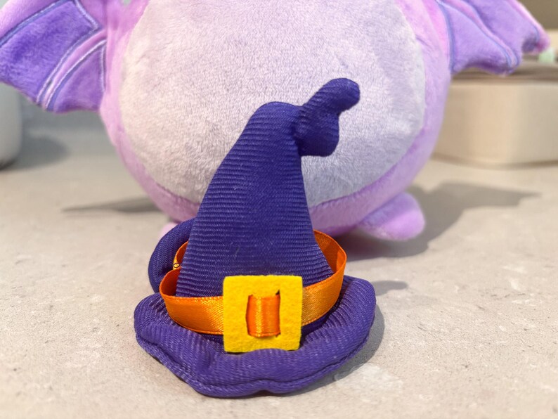 May include: A purple and blue witch hat with an orange ribbon and a yellow buckle. The hat is made of soft fabric and has a pointed top.