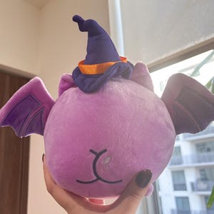 May include: Purple plush bat with a purple and orange witch hat. The bat has a cute face with a small nose and mouth.