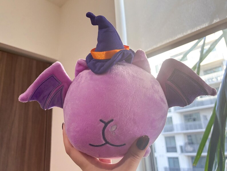 May include: Purple plush bat with a purple and orange witch hat. The bat has a cute face with a small nose and mouth.