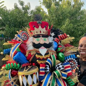 May include: A decorative nutcracker wreath featuring a central nutcracker figure with a red crown, black mustache, and white beard. The wreath is adorned with a miniature drum, multi-colored ribbons in blue, green, red, and black-and-white patterns, and gold foliage accents.
