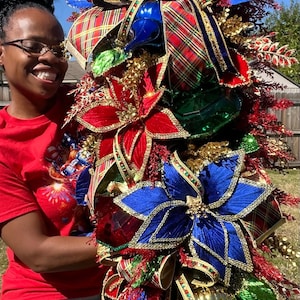 May include: A large decorative Christmas swag featuring red and blue velvet poinsettia flowers, plaid ribbon bows, gold finial ornaments, and red glittered foliage. The arrangement is densely packed with metallic green accents and gold trim.