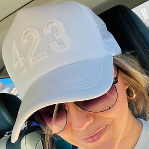May include: White trucker hat with the number "423" embroidered on the front. The hat features a curved brim and a mesh back. The numbers are in a light beige color, contrasting with the white hat.