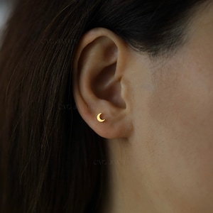 以下が含まれることがあります： 小さな金の三日月型のスタッドピアスをした耳のクローズアップ。ピアスはシンプルで繊細で、毎日の着用に最適です。