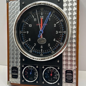 Puede incluir: Un reloj de pared rectangular con esfera negra, borde plateado y marco de madera marrón. El reloj presenta una esfera redonda con las palabras "Spirit of St. Louis" y "Local Time". También incluye dos indicadores más pequeños para la humedad y la temperatura, y una placa con el nombre "Charles Lindbergh".