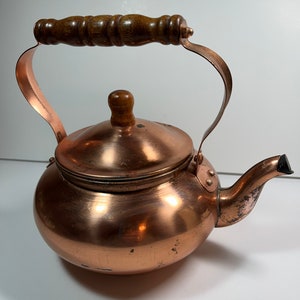 May include: A copper tea kettle with a wooden handle and lid knob. The kettle has a rounded body, a curved handle, and a spout. The copper has a warm, reddish-brown color, and the wood is a dark brown.