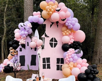 May include: A pink and purple balloon arch with black bats, forming a backdrop for a pink cardboard cutout of a haunted house. Two white chairs and a table with a white tablecloth are in front of the arch.