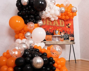 May include: A graduation party decoration featuring a balloon arch with orange, black, white, silver, and clear balloons. The arch is decorated with confetti and is in front of a red and orange backdrop with the word "GRAD" in gold letters.