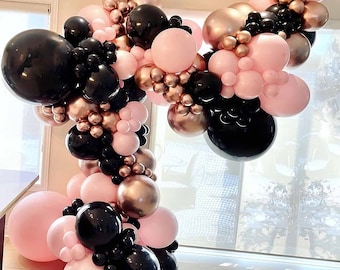 May include: A balloon arch made of black, pink and gold balloons. The arch is made of a variety of sizes and shapes of balloons, and is arranged in a cascading pattern.