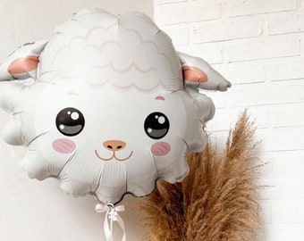 May include: A white balloon shaped like a sheep's head with a pink nose and black eyes. The balloon is tied to a pink ribbon and is in front of a vase of dried pampas grass.