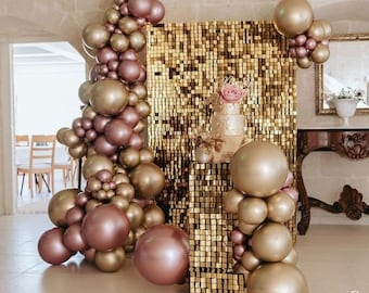 May include: A gold sequin backdrop with a large arrangement of rose gold balloons. The balloons are arranged in a cascading design and are accented with a small bouquet of flowers.