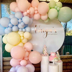 Peut inclure: Des ballons de couleur pastel dans des tons de rose, bleu, jaune et vert sont disposés en forme d'arche. Les ballons sont derrière une pancarte ronde blanche avec le texte "Kaiya's 1st Birthday". Deux piédestaux cylindriques blancs se trouvent au premier plan, l'un avec un gâteau rose décoré de nuages et d'un arc-en-ciel.