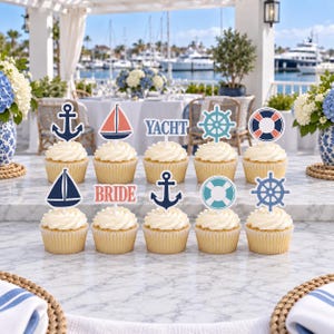 May include: A set of ten cupcakes topped with nautical-themed paper decorations including navy blue anchors, red and white sailboats, a blue ship's wheel, a life preserver, and text cutouts reading YACHT and BRIDE, all arranged on a marble surface.