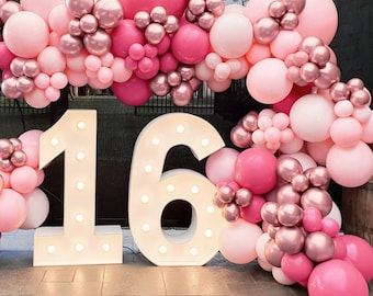 May include: White light-up numbers 16 with a garland of pink and rose gold balloons behind them.