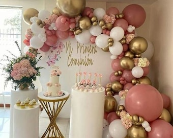 Op de afbeelding: Een witte taart met roze glazuur en gouden accenten staat op een gouden standaard. De taart is omringd door witte, gouden en roze ballonnen. De ballonnen zijn in een boogvorm gerangschikt en zijn versierd met bloemen. De tekst "Mi Primera Comunión" staat op een witte achtergrond achter de taart.