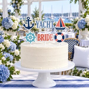 May include: A white cake on a white pedestal stand topped with nautical-themed decorations. The toppers include a dark blue anchor, a light blue ship wheel, a red and blue sailboat, a red and white life preserver, and text reading YACHT BRIDE.
