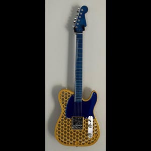 May include: A unique electric guitar with a blue headstock, neck, and pickguard. The body features a honeycomb design in a light brown color. Silver hardware and strings complete the instrument. The guitar is mounted on a white wall.