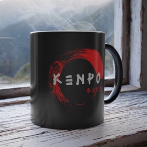 May include: Black ceramic mug with the word "KENPO" in white, inside a red circular design. The mug also has Japanese characters. Steam rises from the mug, suggesting a hot beverage. The mug sits on a weathered, white-painted wooden surface.