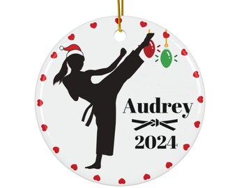 May include: A white ceramic Christmas ornament with a black silhouette of a person performing a martial arts kick, wearing a Santa hat. The ornament has red heart accents and the name "Audrey" with the year "2024".