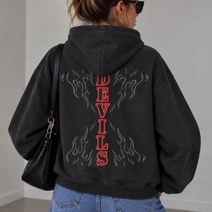 May include: A charcoal gray hooded sweatshirt featuring a vertical red graphic that spells out the word DEVILS in a collegiate font, surrounded by thin white flame outlines on the back of the garment, paired with blue denim jeans and a black bag.
