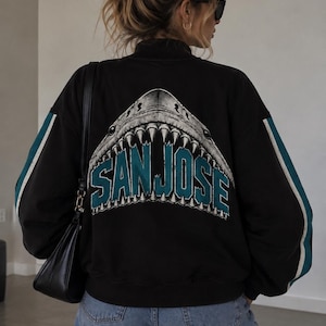 Puede incluir: Una chaqueta negra de manga larga con un gráfico grande de una boca de tiburón con dientes afilados y el texto SAN JOSE en letras verde azulado y blancas en la espalda. Las mangas tienen rayas verde azulado y blancas, cuello alto.