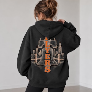 Peut inclure: Un sweat-shirt à capuche noir avec un grand motif au dos. Le design présente le contour clair de la Liberty Bell et une ligne d'horizon urbaine avec un pont, surmontés du mot FLYERS en lettres majuscules orange disposées verticalement.