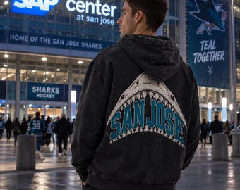 May include: A charcoal grey hooded sweatshirt featuring a large graphic on the back of a shark jaw with the text SAN JOSE in teal and white lettering. The hoodie has a distressed, washed-out appearance and a drawstring hood.