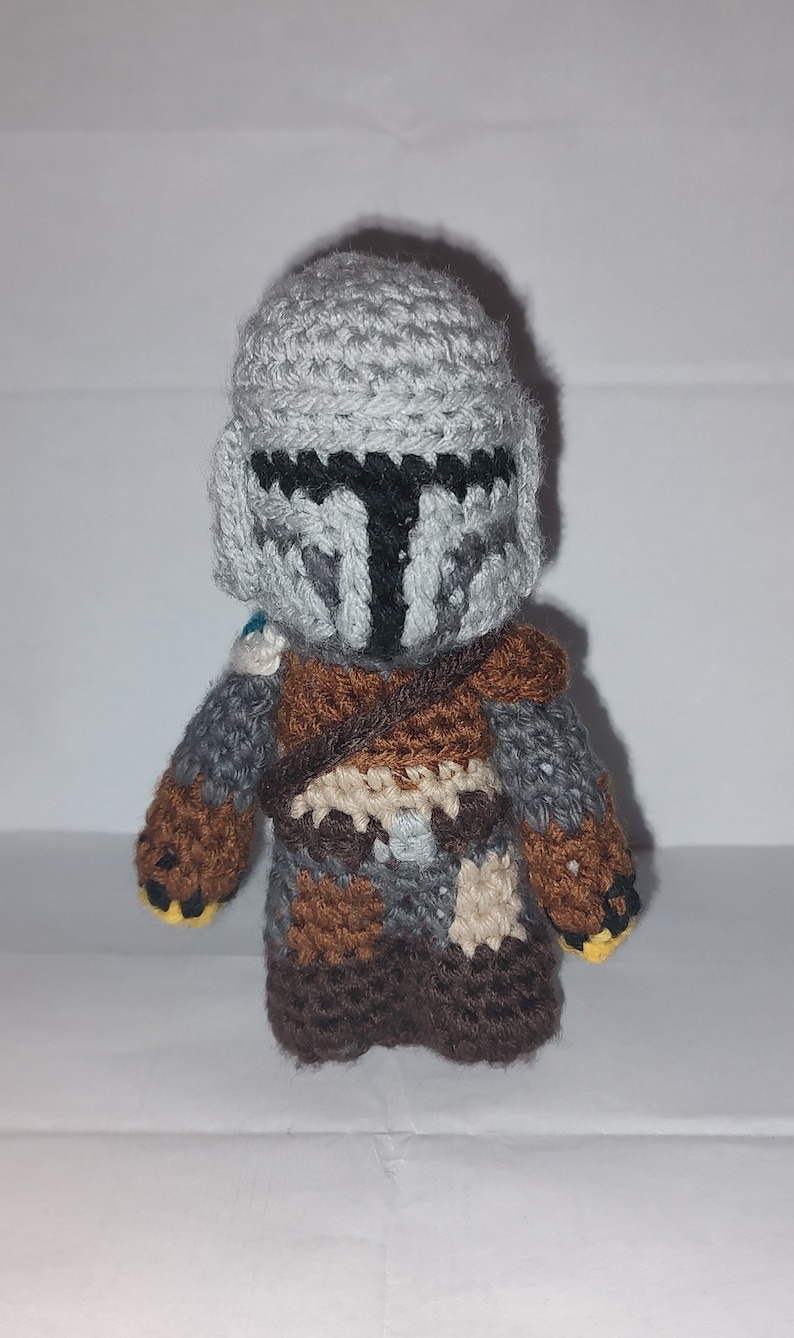 May include: Crocheted Mandalorian figure in gray, brown, and tan. The handmade doll features a helmet with a black T-shaped visor, brown shoulder armor, and a brown and tan outfit. The figure has yellow accents on the hands.