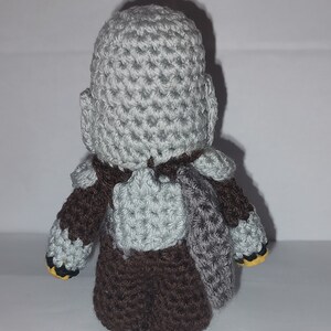 May include: A handmade crochet figure of a character, viewed from the back. The figure has a gray head and shoulders, brown torso and arms, and gray armor-like details. The figure's feet have yellow accents.