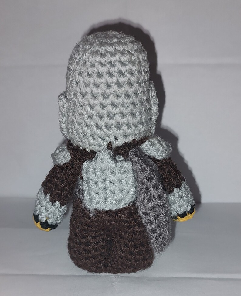 May include: A handmade crochet figure of a character, viewed from the back. The figure has a gray head and shoulders, brown torso and arms, and gray armor-like details. The figure's feet have yellow accents.