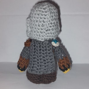 May include: A handmade crochet doll with a gray helmet and body, brown arms, and a dark brown base. The doll has small details like a blue and white accent on the shoulder and yellow accents on the hands. The doll is made of yarn.