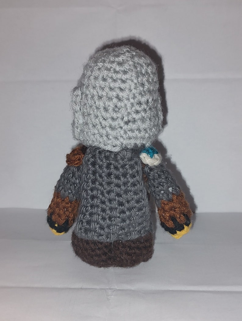 May include: A handmade crochet doll with a gray helmet and body, brown arms, and a dark brown base. The doll has small details like a blue and white accent on the shoulder and yellow accents on the hands. The doll is made of yarn.