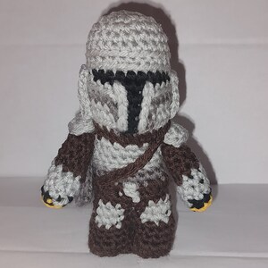 May include: A handmade crochet figure of a character from a popular science fiction series. The figure is primarily gray, brown, and black, with a helmet and armor. The figure has a brown strap across the chest and yellow accents on the hands.