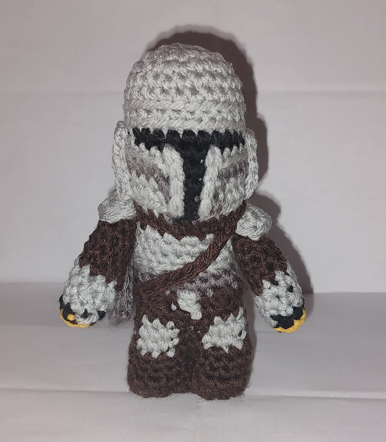 May include: A handmade crochet figure of a character from a popular science fiction series. The figure is primarily gray, brown, and black, with a helmet and armor. The figure has a brown strap across the chest and yellow accents on the hands.